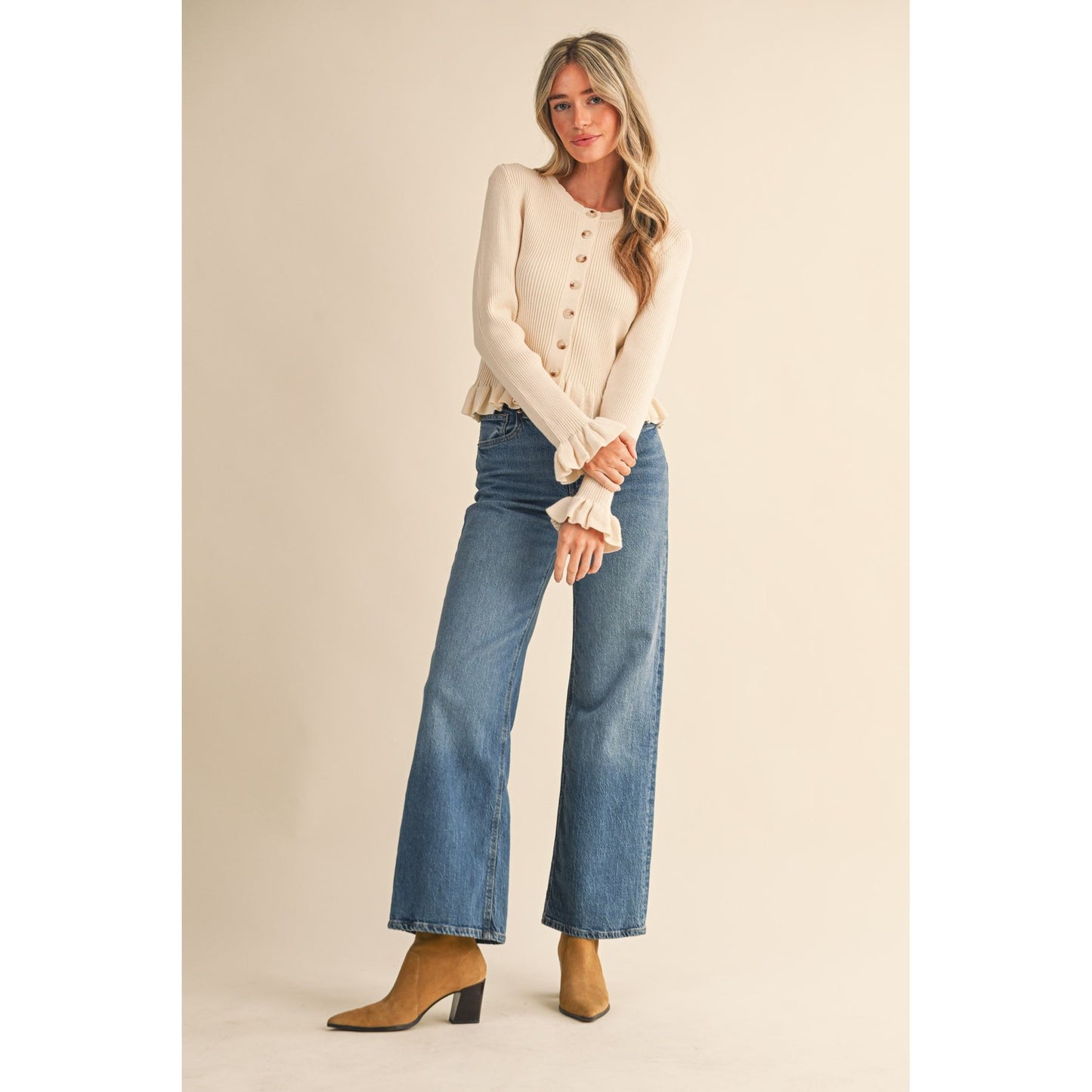 Woman wearing a cream cardigan, blue jeans, and brown boots on a beige background