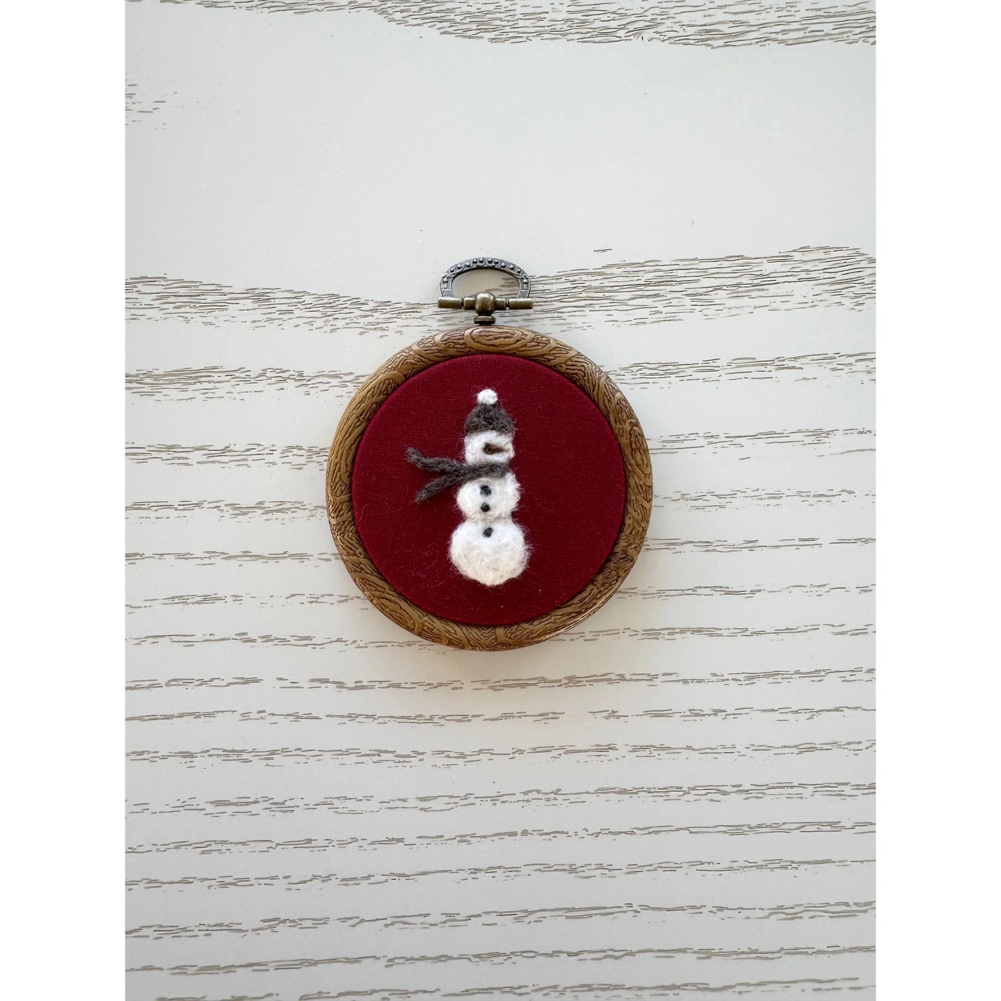 Embroidery hoop with a snowman design on a red background, placed on a textured white surface.