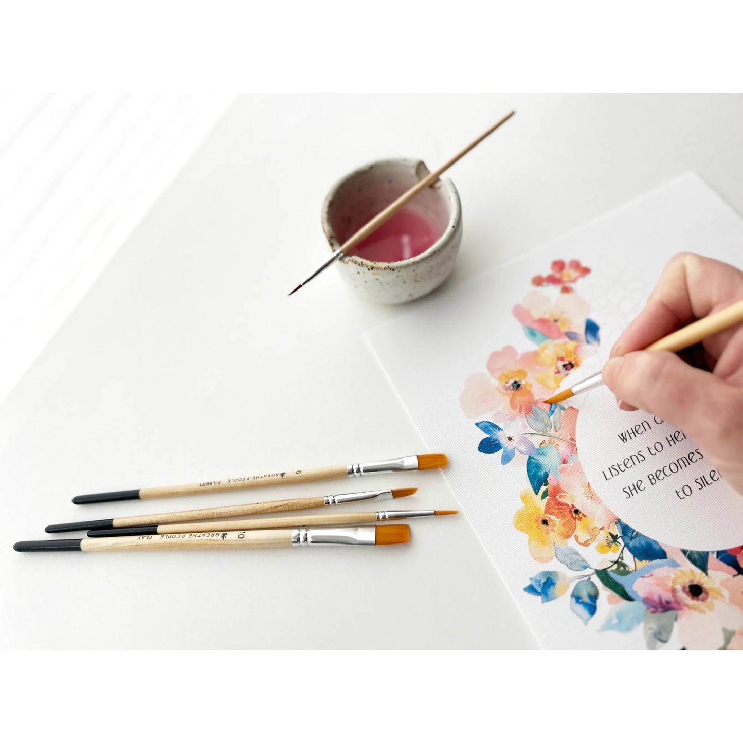 Person painting a floral design on paper with brushes and a small container of paint.