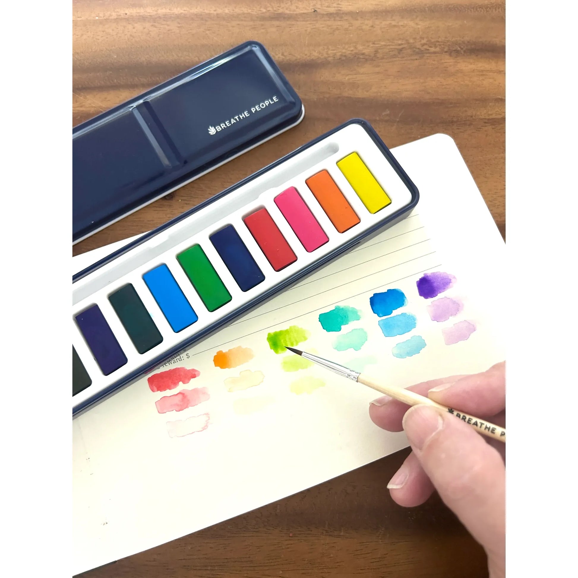 Watercolor paint set with a hand holding a brush and painting on paper.