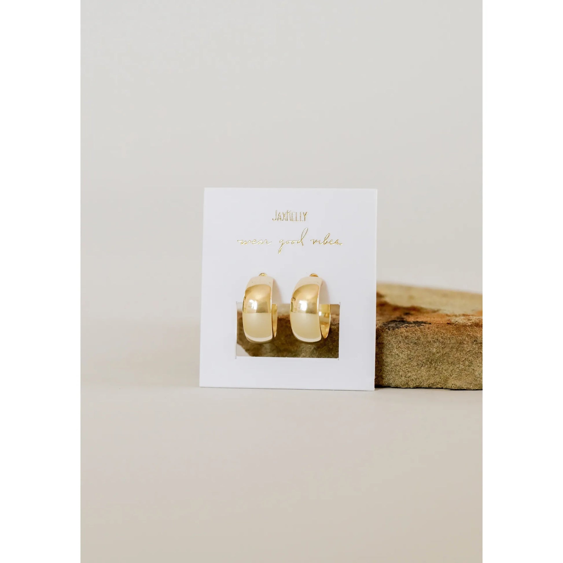 Gold hoop earrings on a white card with gold text, placed on a wooden block against a beige background.