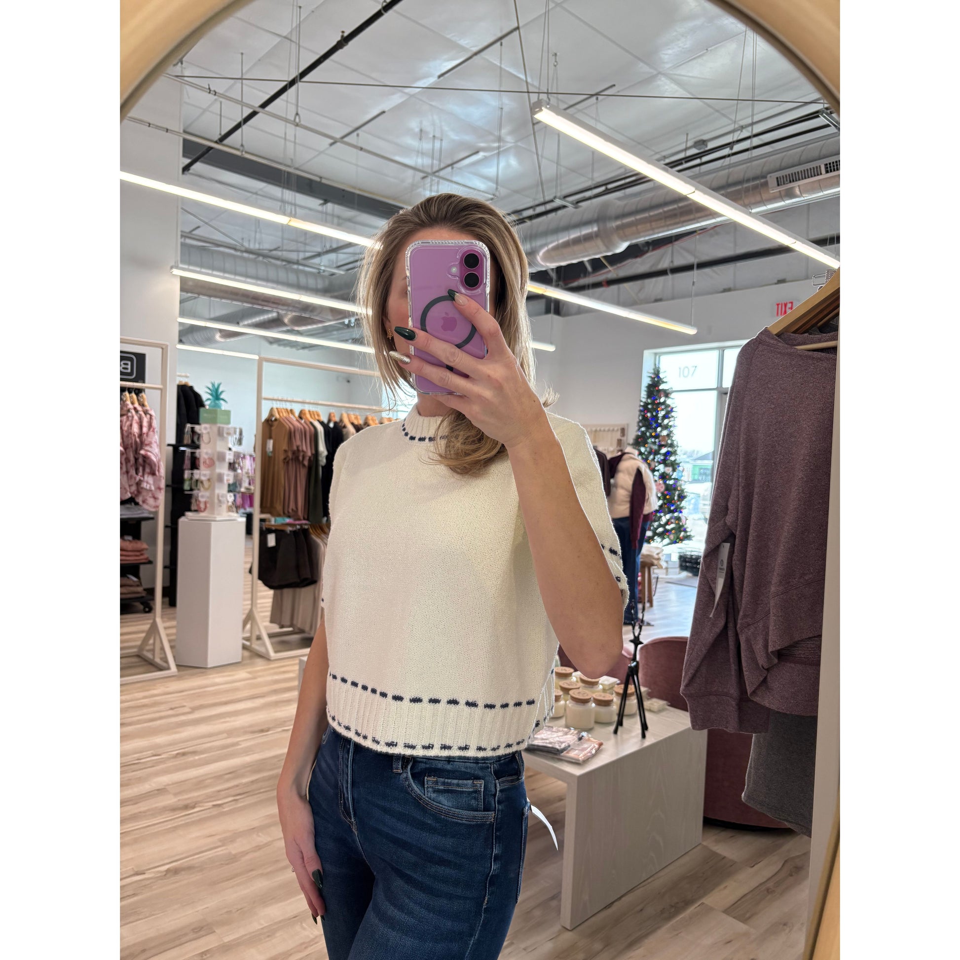 Person taking a mirror selfie in a store, wearing a cream short-sleeve knit sweater