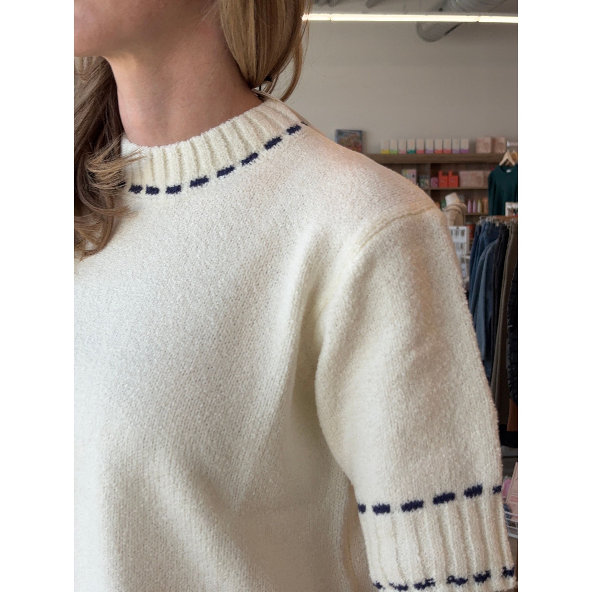 Close-up of a person wearing a cream sweater with navy stitching details in a store setting