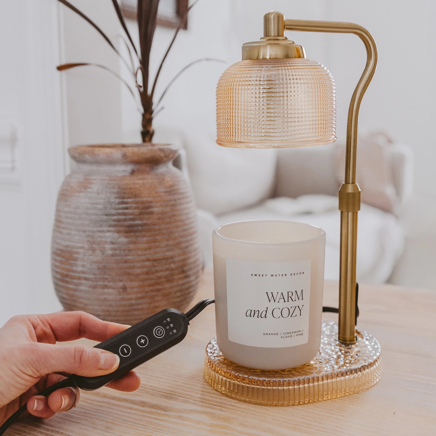 Hand holding a remote control next to a candle labeled 'Warm and Cozy' on a table with a lamp in the background.