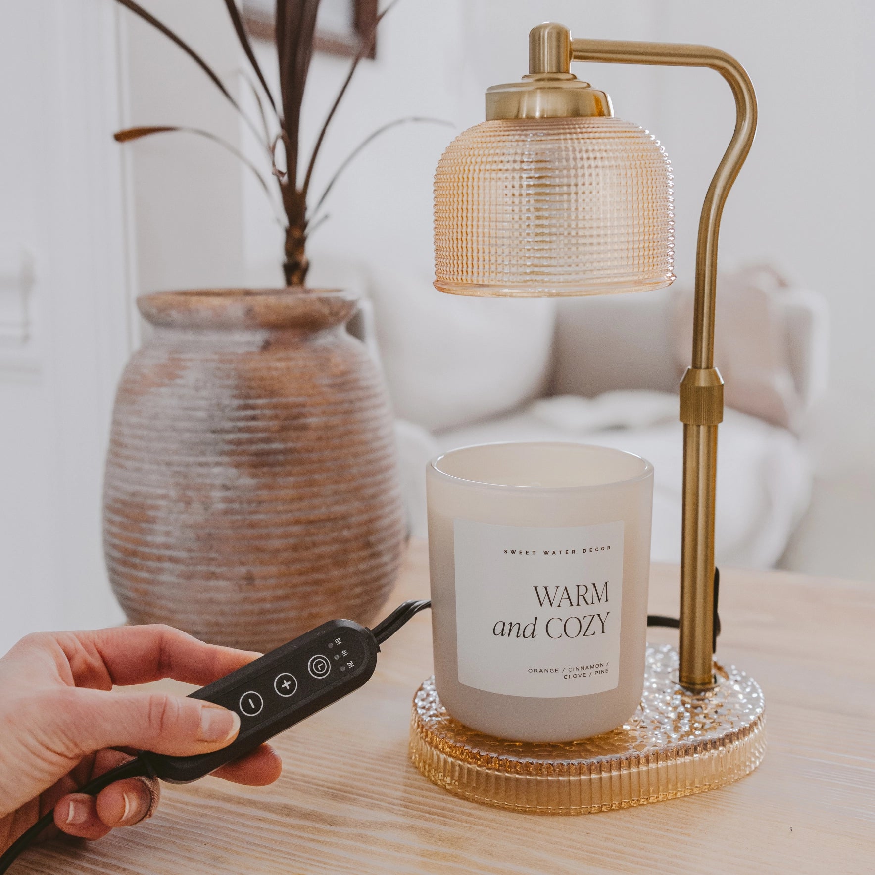 Hand holding a remote control next to a candle labeled 'Warm and Cozy' on a table with a lamp in the background.
