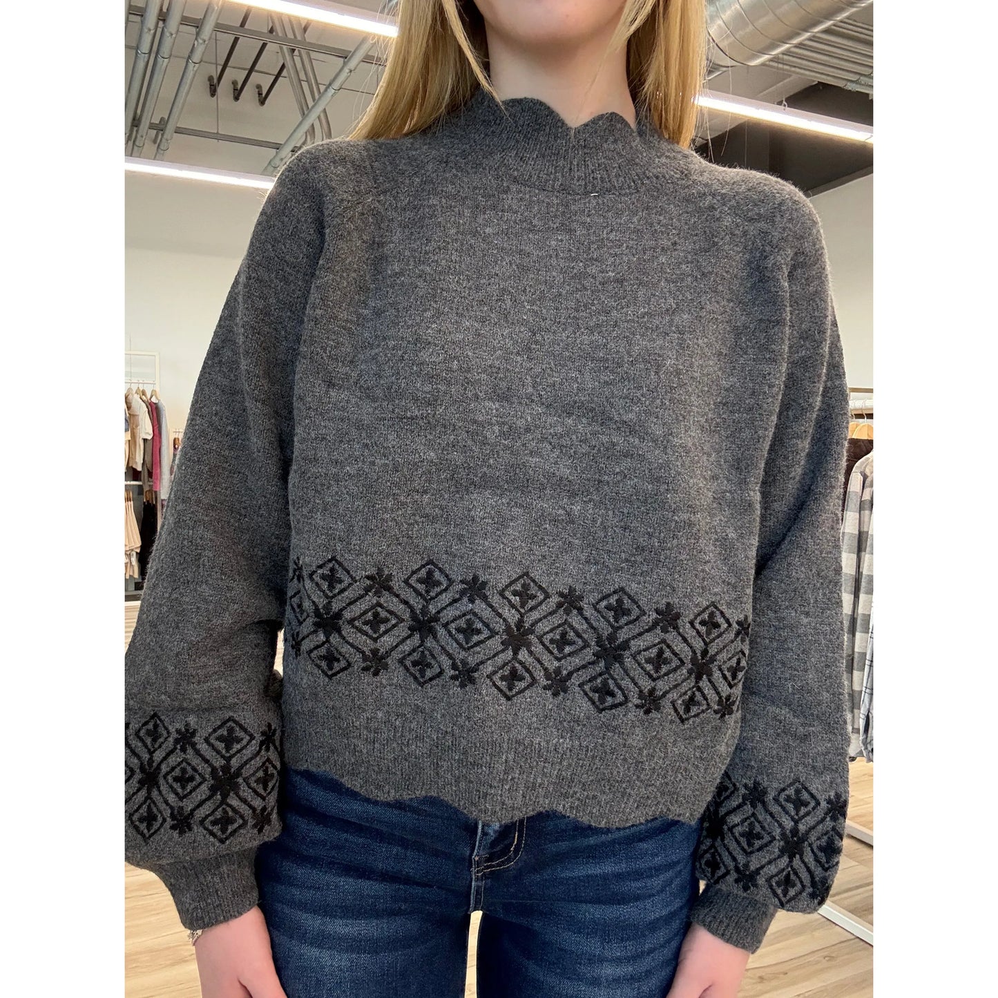 Person wearing a gray sweater with a black embroidered pattern in a store setting