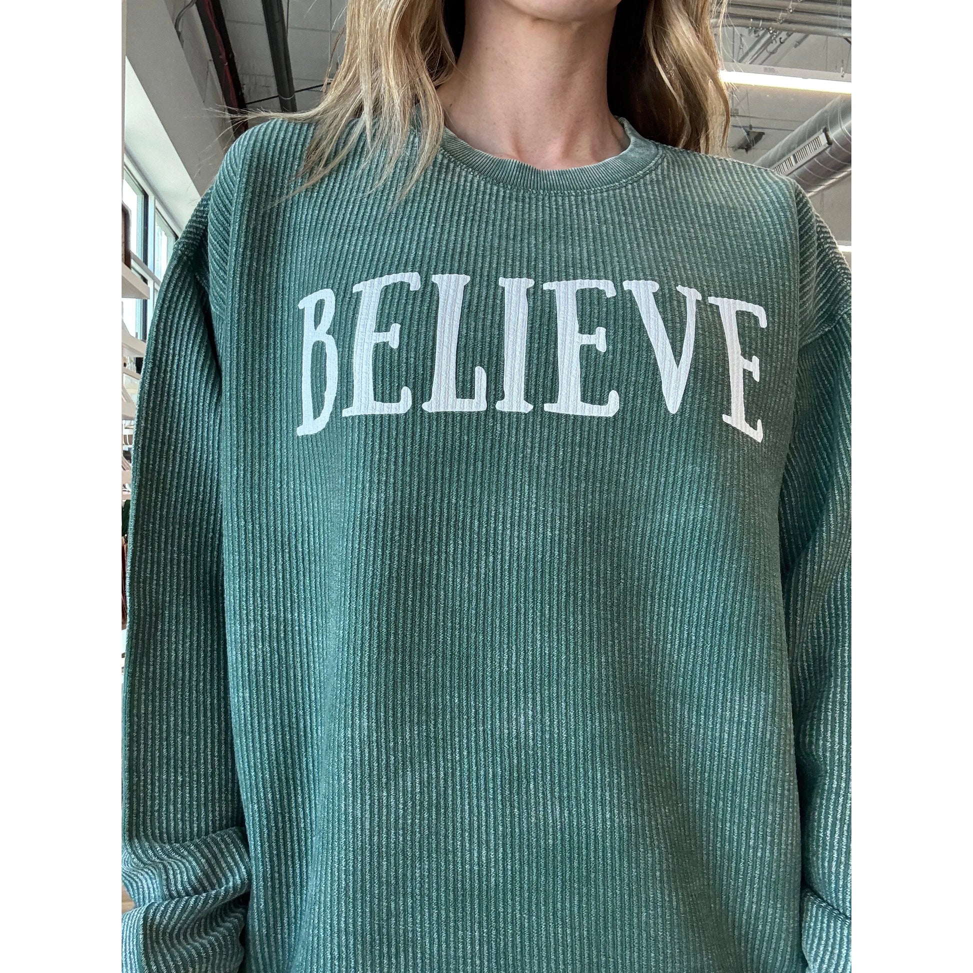 Green ribbed sweater with 'BELIEVE' text worn by a person indoors.