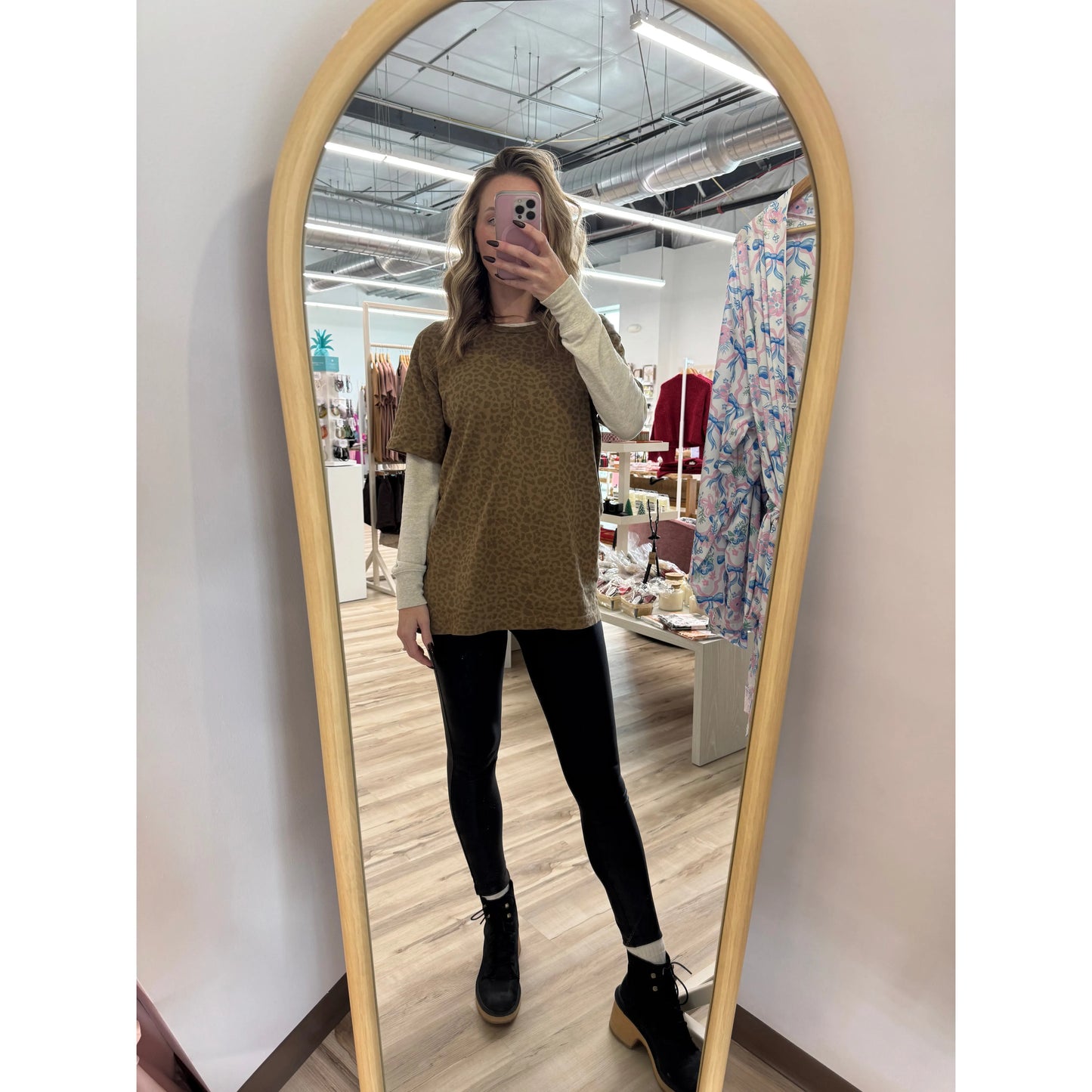 Person taking a mirror selfie wearing a brown leopard shirt and black pants in a store.