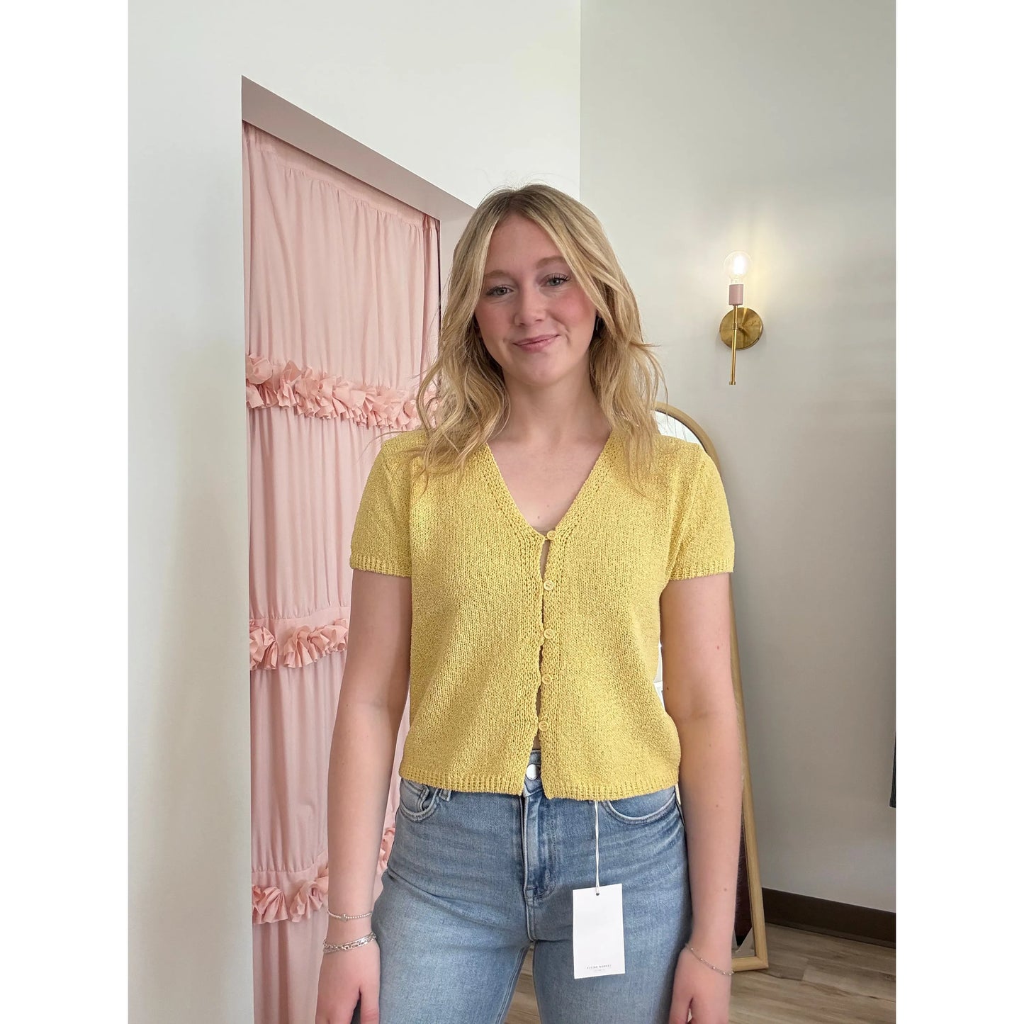 Woman wearing a yellow short-sleeve cardigan and blue jeans standing in a room with pink curtains.