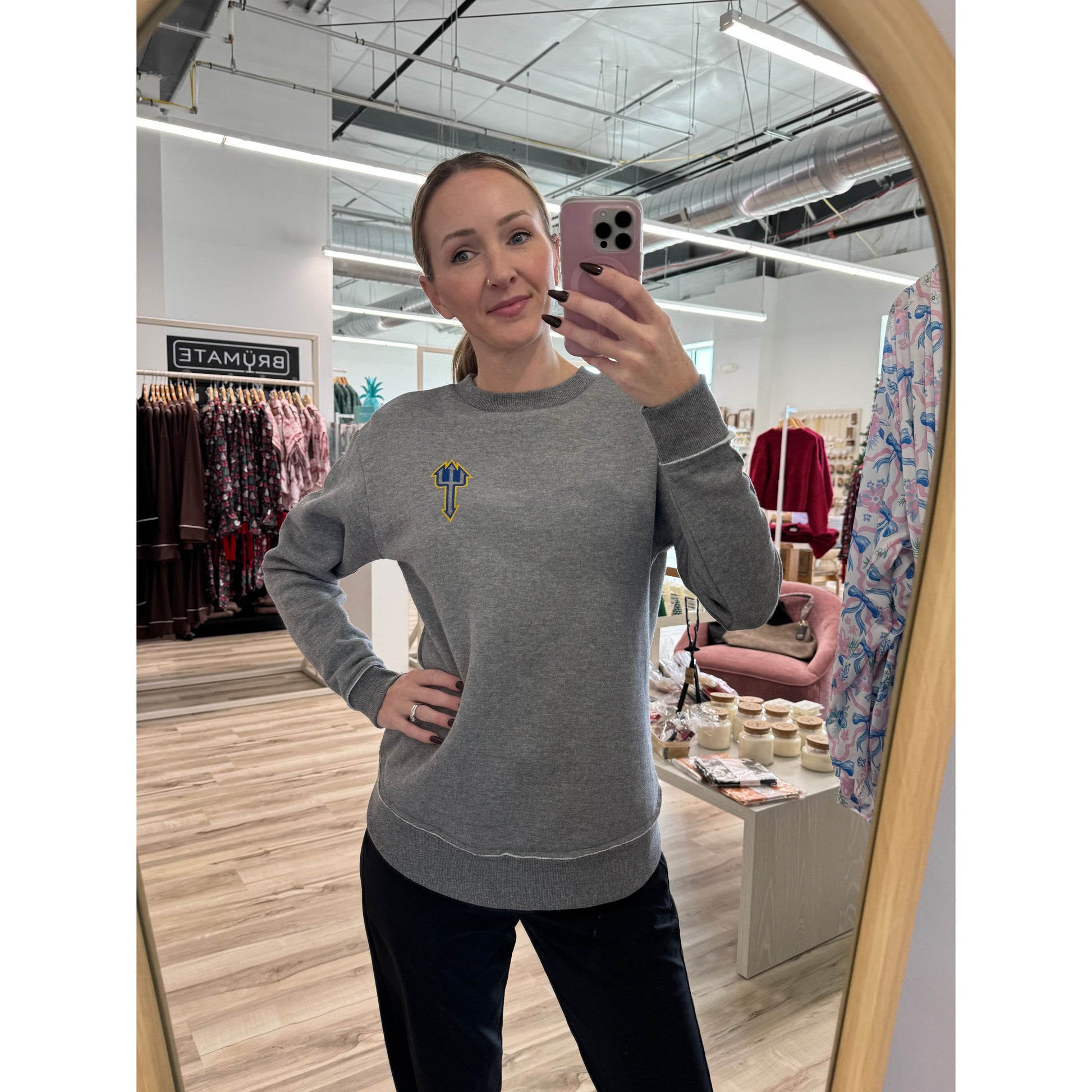 Person taking a mirror selfie wearing a gray sweatshirt with a logo in a store setting.