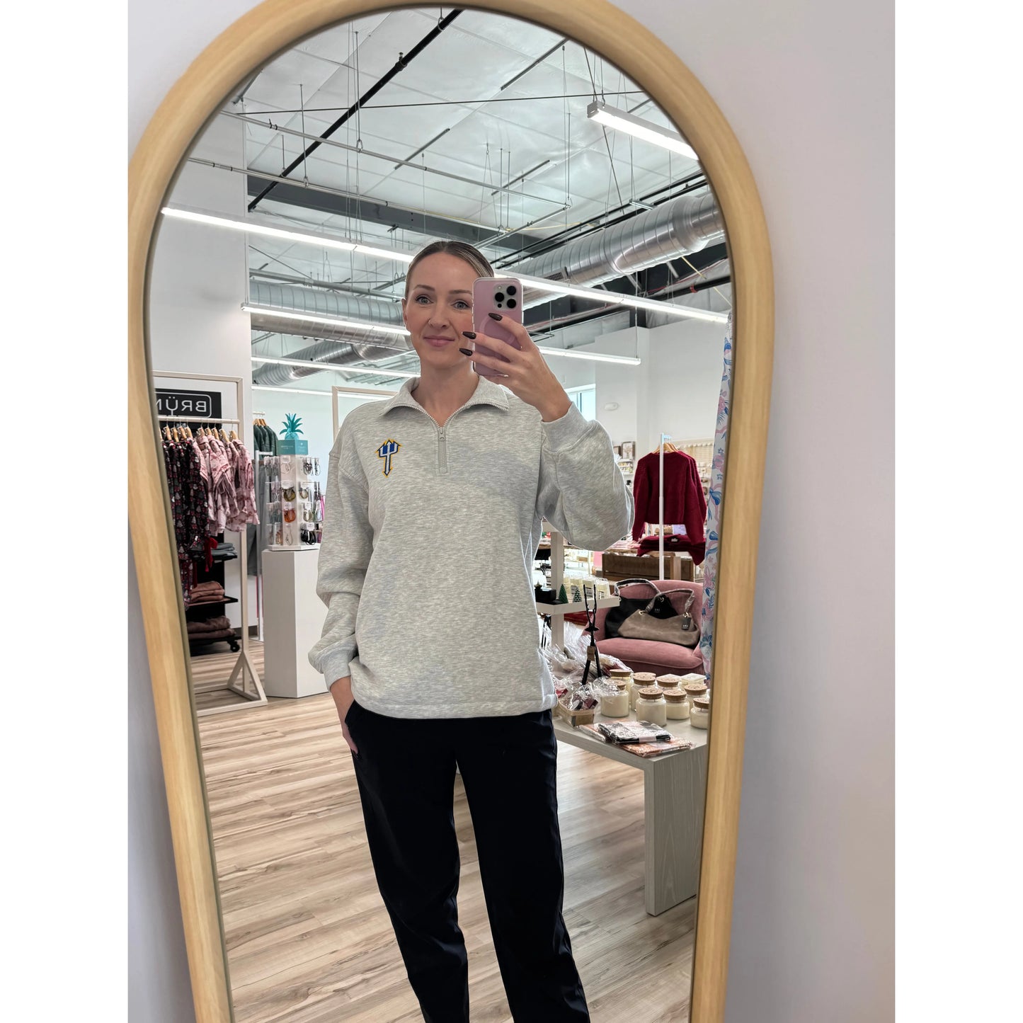 Person taking a mirror selfie wearing a light gray sweater with a logo in a store setting.
