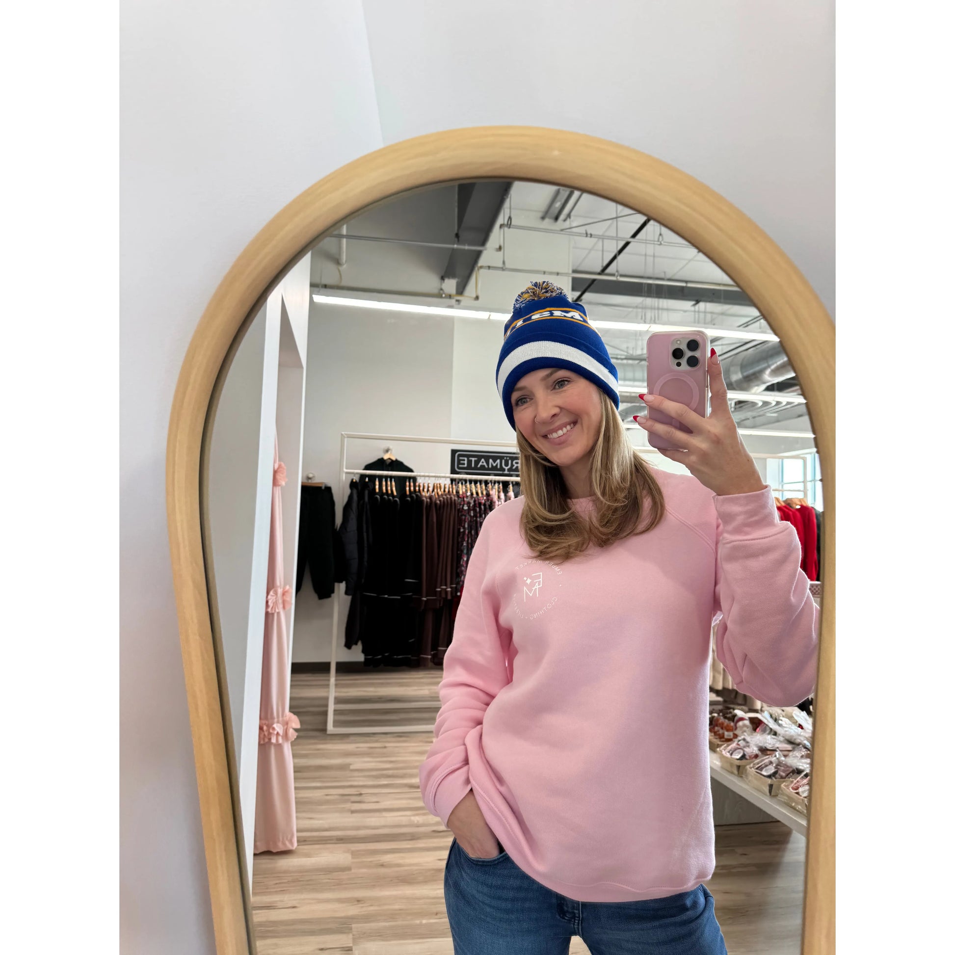 Person wearing a pink sweatshirt and blue knit beanie, taking a mirror selfie in a store.