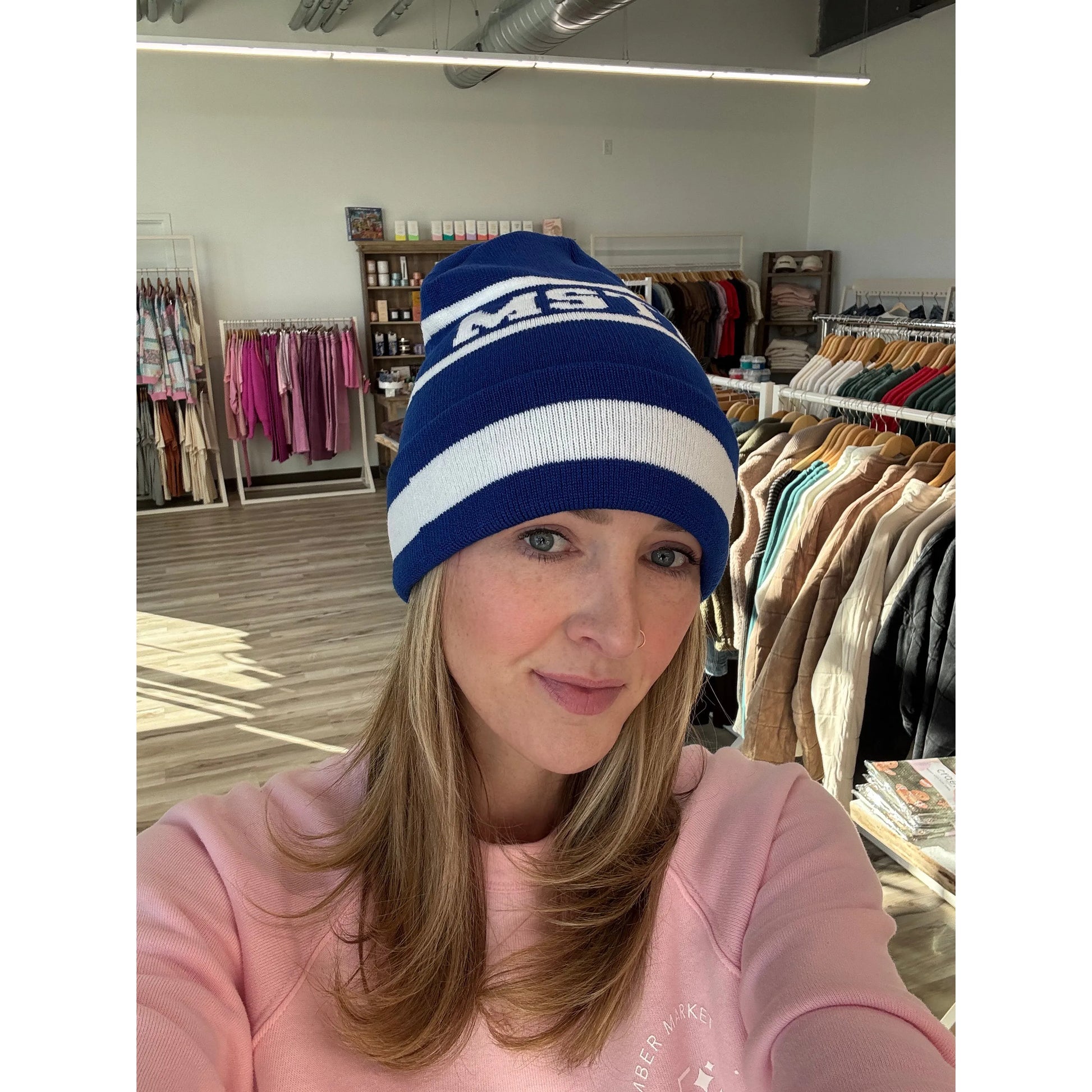 Woman wearing a blue and white beanie with 'MSTM' logo in a clothing store.