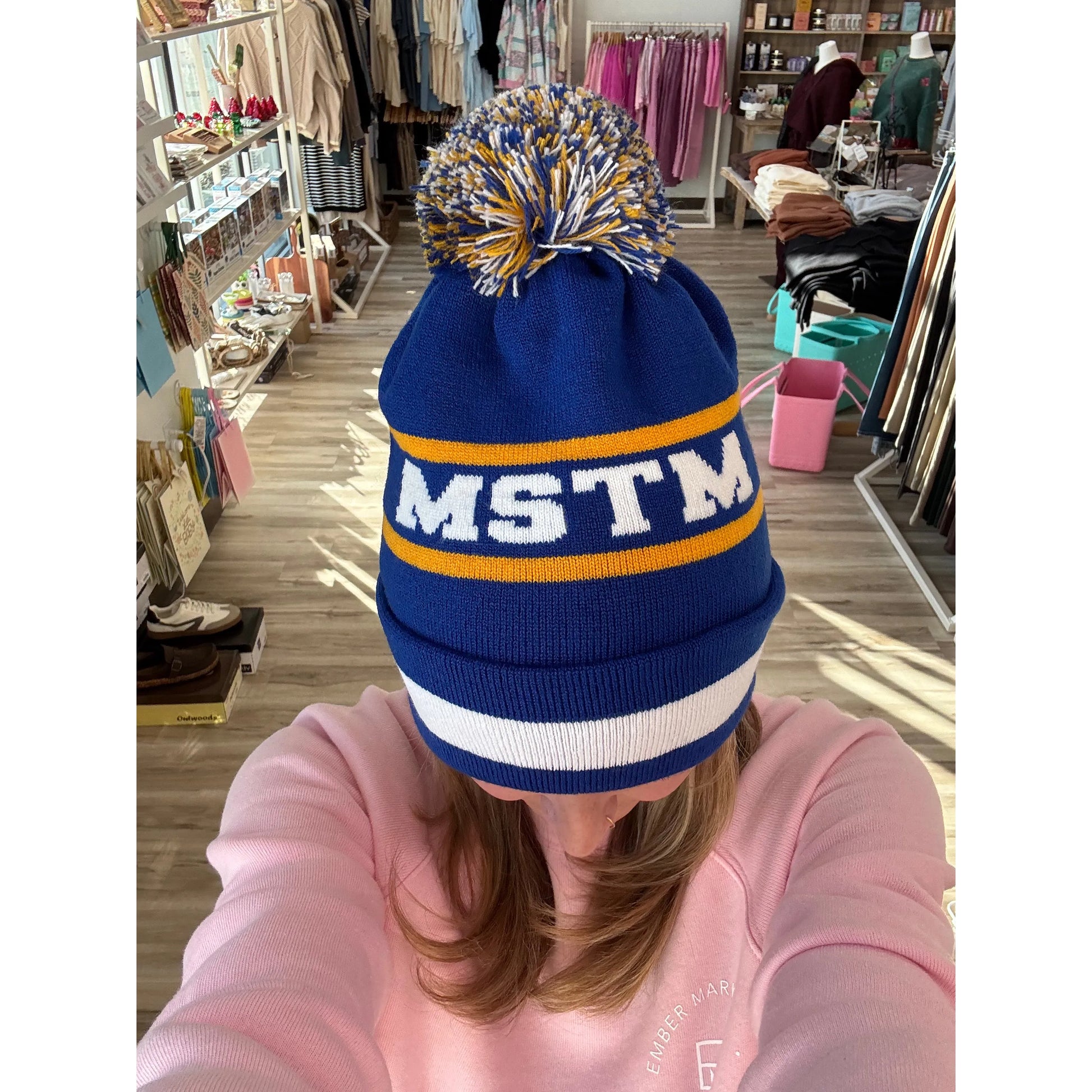 Person wearing a blue beanie with pom-pom with 'MSTM' on it in a store setting