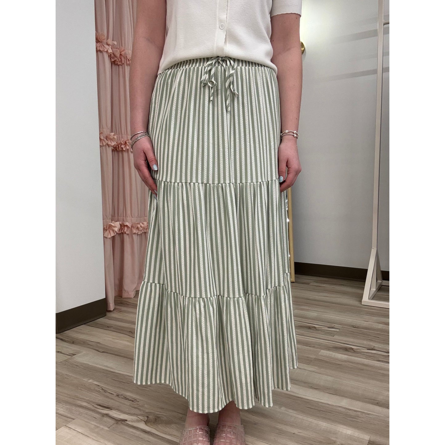 Person wearing a white shirt and green striped skirt in a room with wooden flooring and a pink curtain.
