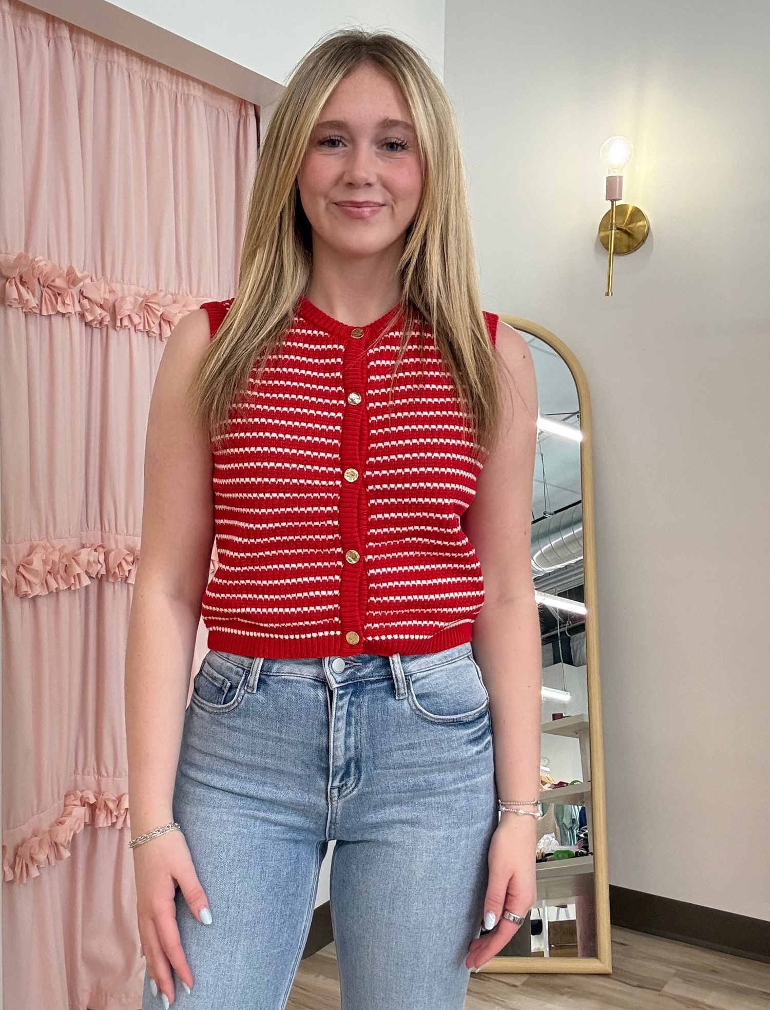Woman wearing a red top with white patterns and blue jeans standing in a room with a mirror and pink curtain.