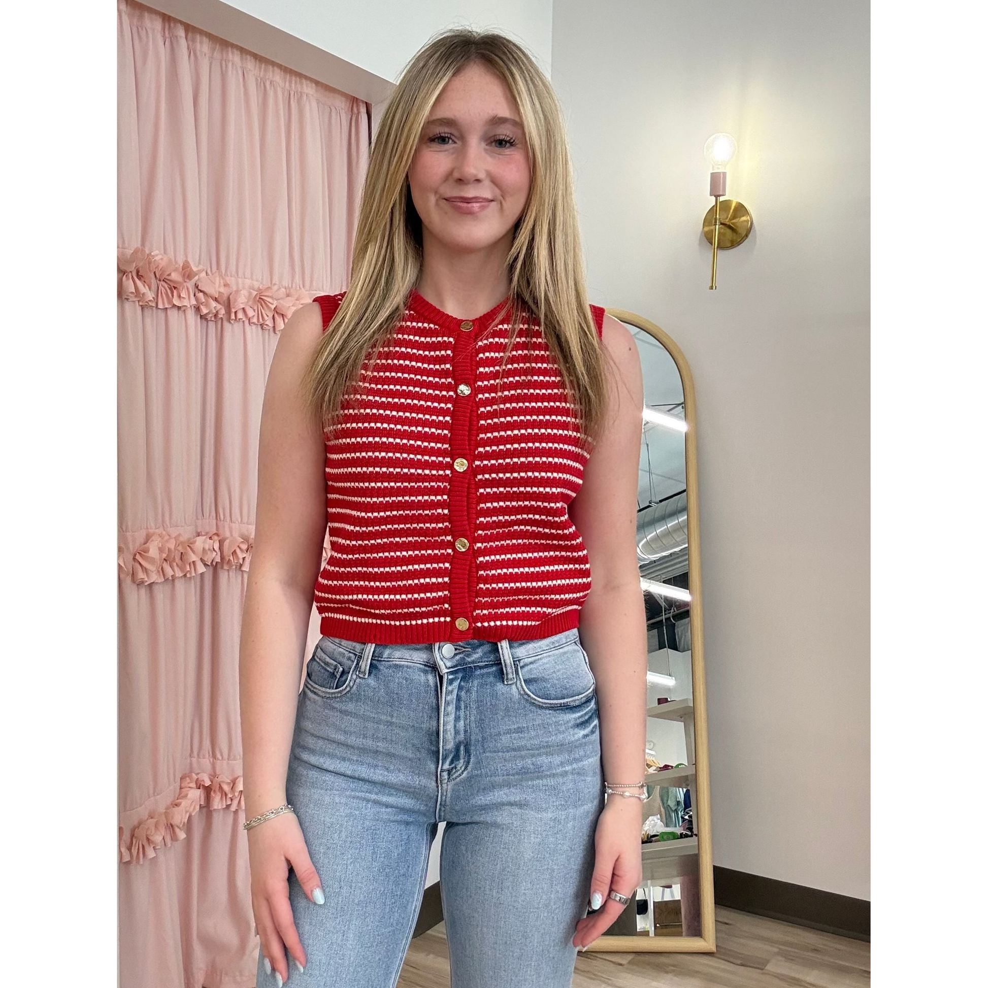 Woman wearing a red top with white patterns and blue jeans standing in a room with a mirror and pink curtain.