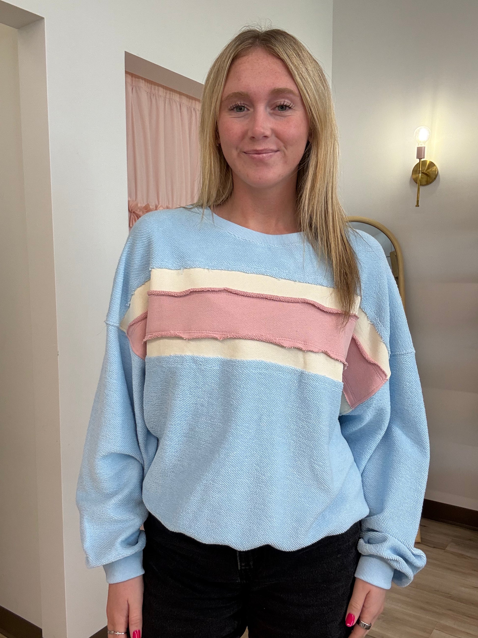 A woman with blonde hair wearing a light blue crewneck sweatshirt with horizontal stripes in cream and pink across the chest, paired with black pants, standing in an indoor setting.