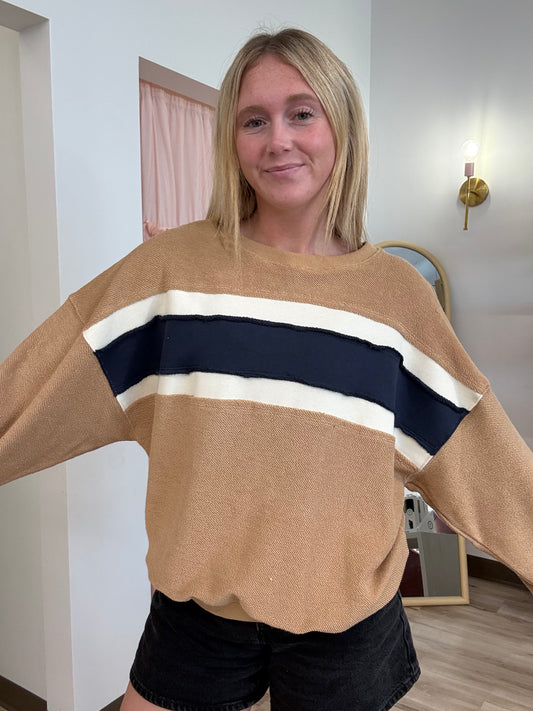 A woman wearing a beige knit sweater with a wide navy blue and cream stripe across the chest, paired with black denim shorts, standing in an indoor room.