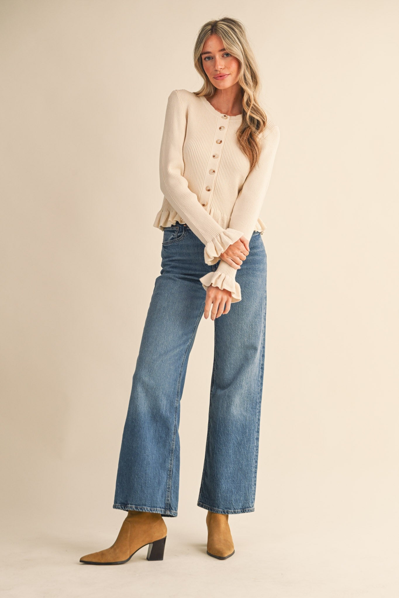 Woman wearing a cream cardigan, blue jeans, and brown boots on a beige background