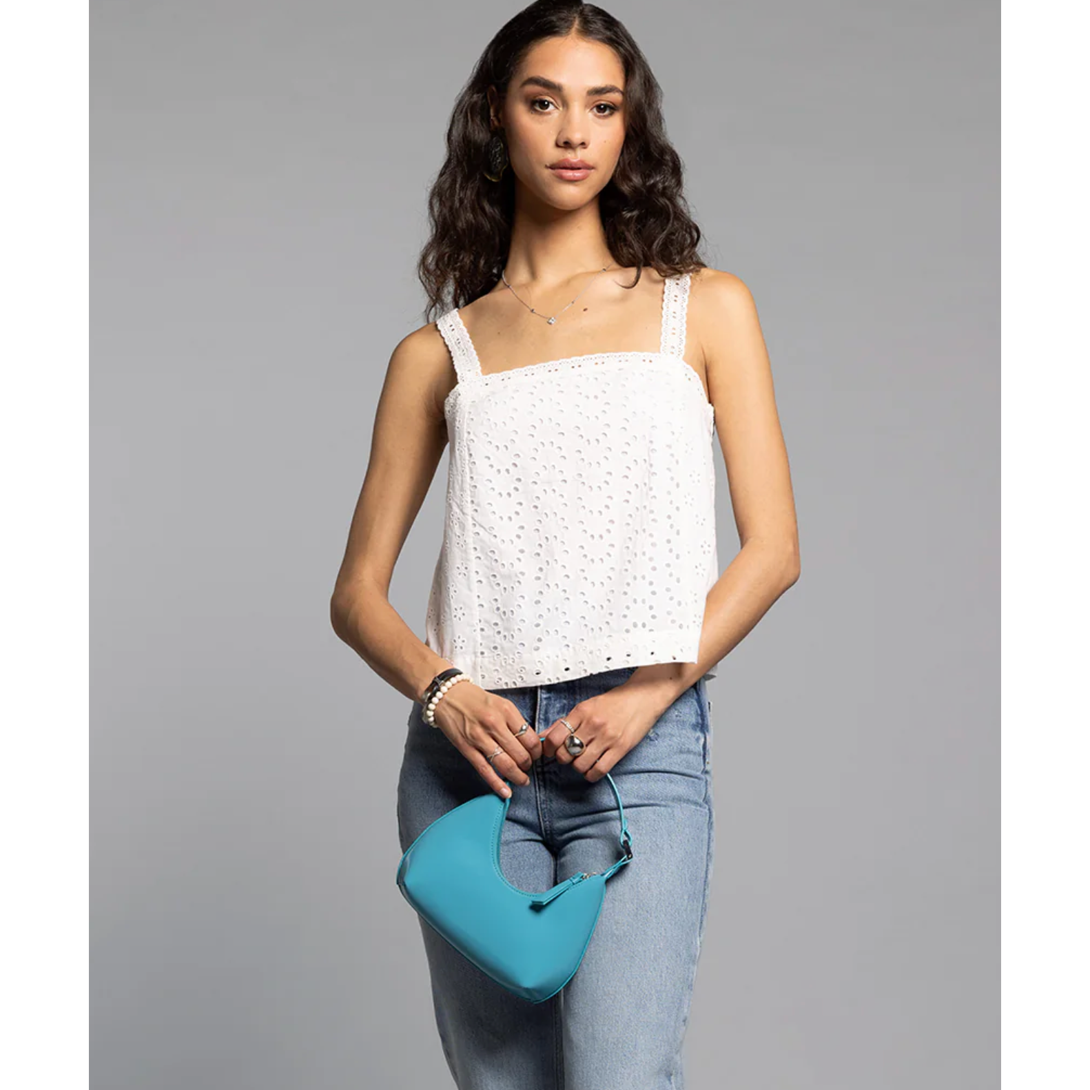 Woman wearing a white top and blue jeans, holding a blue handbag against a gray background