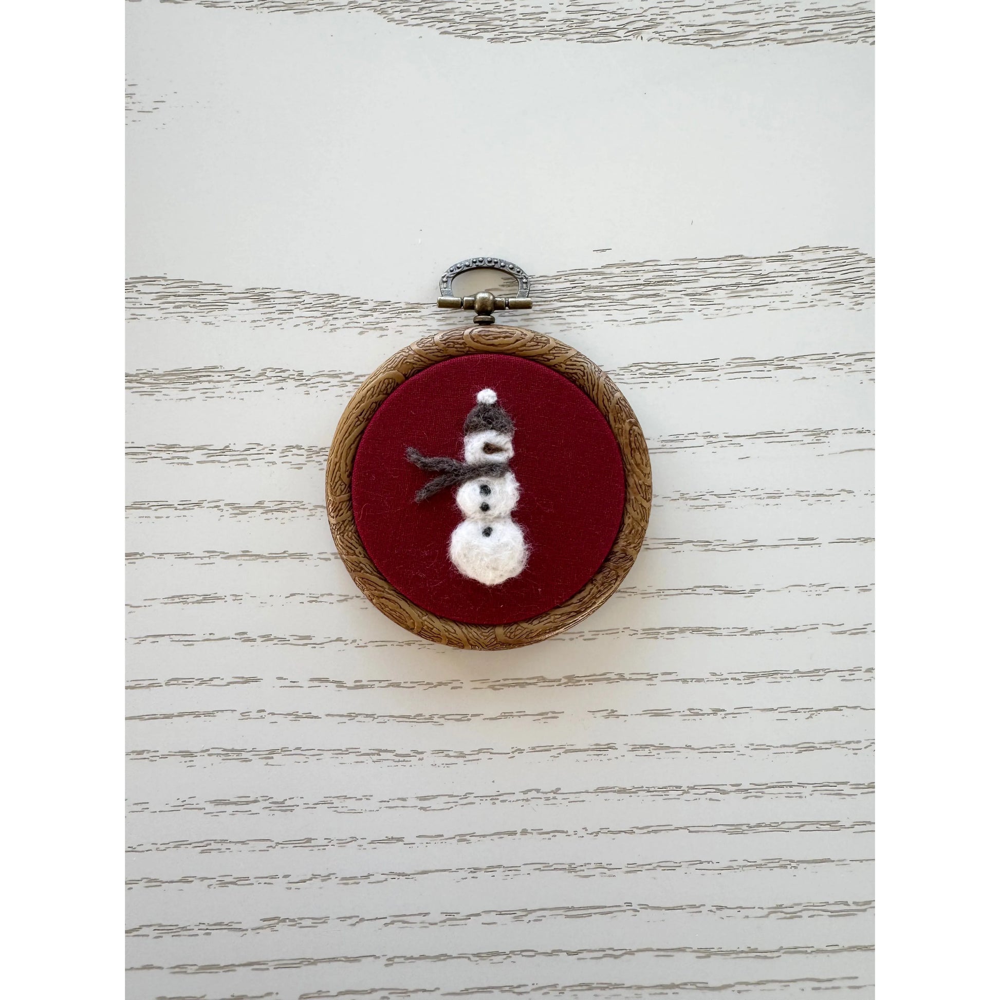 Embroidery hoop with a snowman design on a red background, placed on a textured white surface.