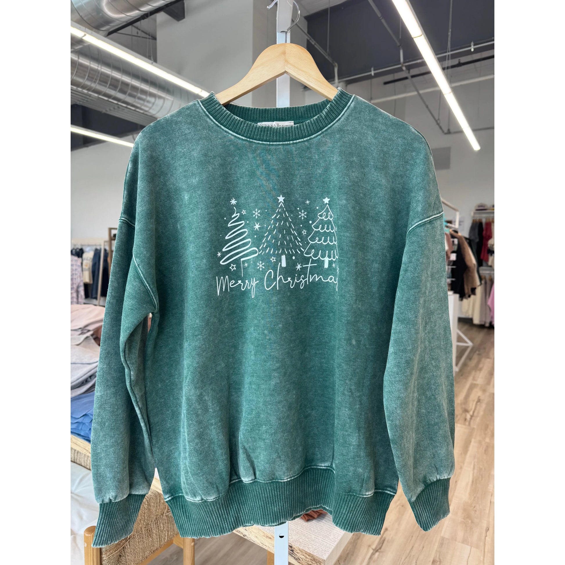 Green sweater with Christmas tree design and 'Merry Christmas' text on a hanger in a store.